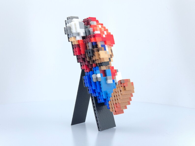 May include: A pixelated figure of Mario from the Super Mario Bros. video game series. The figure is made of small plastic blocks and is standing on a black stand. The figure is wearing a red hat, blue overalls, and white gloves.