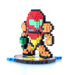 May include: A pixelated figure of a video game character, Samus Aran from the Metroid series, made of colorful plastic blocks. The figure is standing on a black base with a checkered pattern.