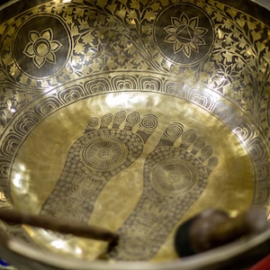 Puede incluir: Un cuenco cantón dorado con diseños intrincados y dos huellas de pies estilizadas en el interior. El cuenco se golpea con un mazo de madera.