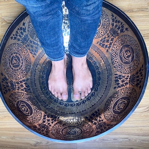 Puede incluir: Los pies descalzos de una persona están colocados dentro de un cuenco de canto grande, redondo y de cobre con intrincados diseños y símbolos. El cuenco tiene un borde negro y está decorado con un patrón de color cobre.
