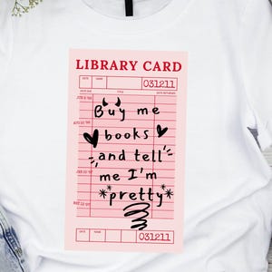 May include: White t-shirt with a pink library card design. The card reads "Buy me books, and tell me I'm pretty" in black script. The shirt is paired with blue jeans, white sneakers, and black headphones.