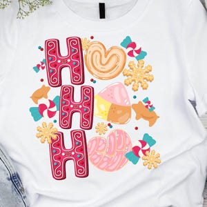 May include: White t-shirt featuring a festive "HO HO HO" design in red gingerbread-style lettering. The design includes gingerbread cookies, candies, and snowflakes in various colours. The shirt is paired with blue jeans, white trainers, and black headphones.