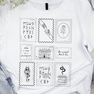 Puede incluir: Camiseta blanca con diseños de sellos postales en blanco y negro. Los sellos muestran manos, una rosa, un edificio, una serpiente y textos como "Like a rose in a graveyard" y "Be careful. Don't die."