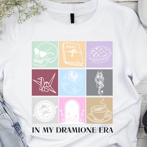 May include: White t-shirt with a colorful grid design featuring various illustrations, including a skull, book, and pie. The text "IN MY DRAMIONE ERA" is printed at the bottom. The shirt is displayed with jeans, sneakers, and headphones.