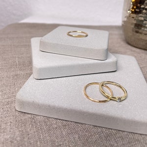 Könnte beinhalten: Drei weiße quadratische Beton-Ringteller mit drei goldenen Ringen darauf. Die Ringe sind schlicht und zart.