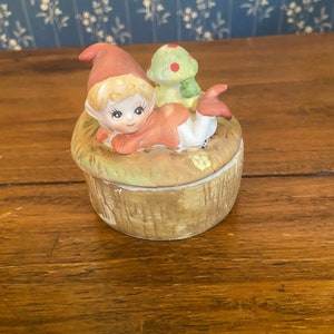 May include: A ceramic trinket box featuring a whimsical design. The box is shaped like a mushroom and adorned with a reclining gnome figure wearing a red hat and a small green mushroom with red spots. The box is painted in earthy tones.