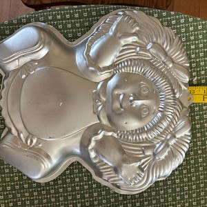 May include: Silver-colored cake pan in the shape of a woman's face with a bonnet and bow. The pan measures approximately 16 inches in length. The text "FOR BEAUTY" is visible on a yellow measuring tape.