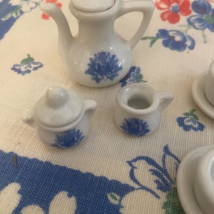 May include: A miniature white porcelain tea set with blue floral designs. The set includes a teapot, a sugar bowl, and a creamer. The pieces are arranged on a floral patterned tablecloth.