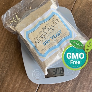 May include: A clear plastic bag containing 300 grams of dry yeast on a digital kitchen scale. The bag has a blue and white label that reads "The French Bakery Est. 1984 Dry Yeast" and "Instant Dry Yeast 300 g/11 oz - Product of France Best if used by 10/2021 - www.ThefrenchBakeryUS.com". A green circle with the text "GMO Free" is visible in the upper right corner of the image.