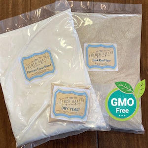 May include: Three white plastic bags containing baking ingredients: Parisian Flour Blend, Dark Rye Flour, and Dry Yeast. The bags are on a wooden surface. A green and white sticker with the text "GMO Free" is visible.
