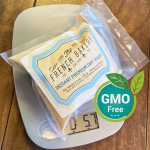 May include: A white digital kitchen scale with a clear plastic bag containing a beige powder labeled "The French Bakery Instant Premium Dry Yeast" with a net weight of 5.3 oz / 150 gr. The scale reads 5.7 oz. A green circle with the text "GMO Free" is in the lower right corner of the image.