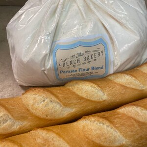 May include: A large white bag of The French Bakery Parisian Flour Blend, next to two long, golden brown baguettes.