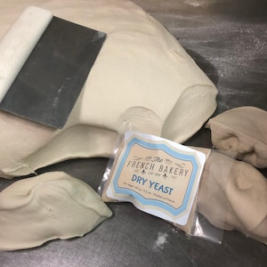 May include: A close-up of a white dough with a metal dough scraper. The dough is divided into four pieces. A small white bag with a label that reads "The French Bakery Est. 1984 Dry Yeast Dry Yeast 100 g / 3.5 oz. - Product of France" is in the center of the image.