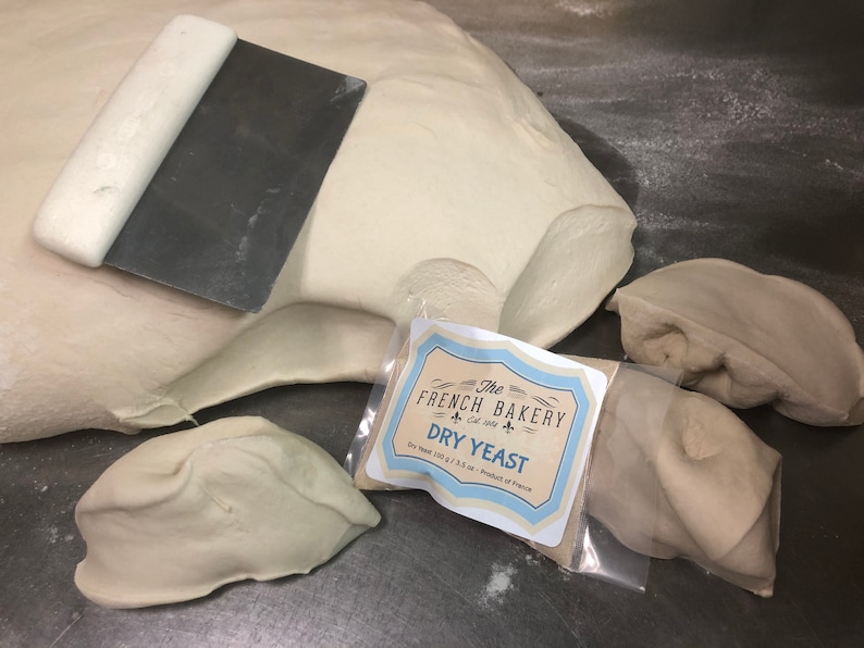 May include: A close-up of a white dough with a metal dough scraper. The dough is divided into four pieces. A small white bag with a label that reads "The French Bakery Est. 1984 Dry Yeast Dry Yeast 100 g / 3.5 oz. - Product of France" is in the center of the image.