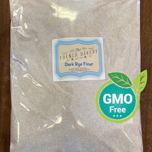 May include: A clear plastic bag filled with dark rye flour. The bag has a label that reads "The French Bakery" and "Dark Rye Flour." A green and white "GMO Free" sticker is on the bag. The bag is sitting on a wooden surface.