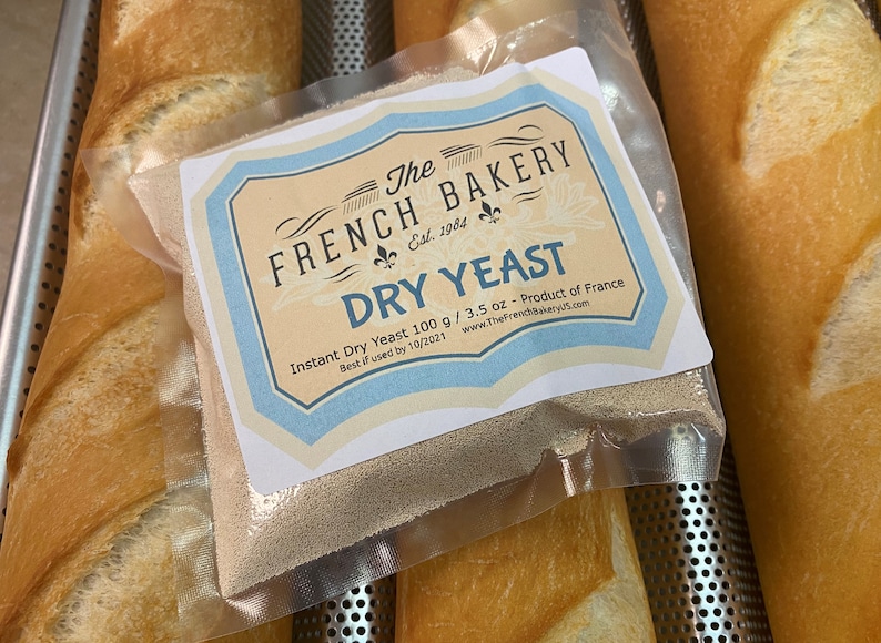 May include: A white bag of dry yeast with a blue and white label that reads "The French Bakery Est. 1984 Dry Yeast Instant Dry Yeast 100 g / 3.5 oz - Product of France Best if used by 10/2021 www.TheFrenchBakeryUS.com". The bag is sitting on top of a loaf of bread.