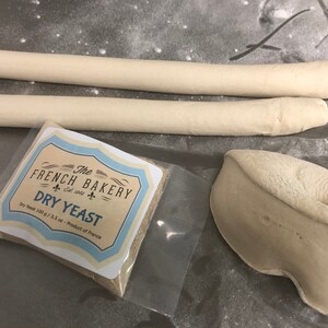 May include: A close-up of a white plastic bag containing a small packet of dry yeast. The packet is labeled "The French Bakery Est. 1965 Dry Yeast" and "Dry Yeast 100 g / 3.5 oz - Product of France".