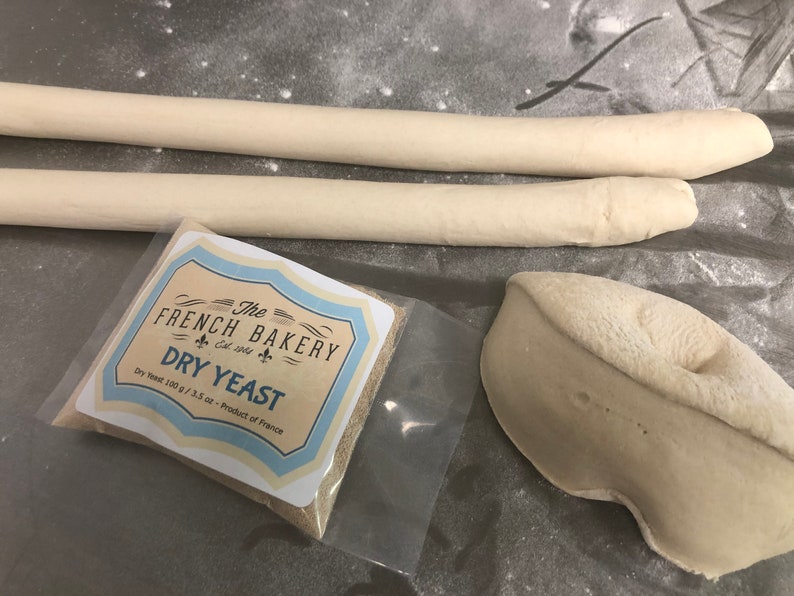 May include: A close-up of a white plastic bag containing a small packet of dry yeast. The packet is labeled "The French Bakery Est. 1965 Dry Yeast" and "Dry Yeast 100 g / 3.5 oz - Product of France".