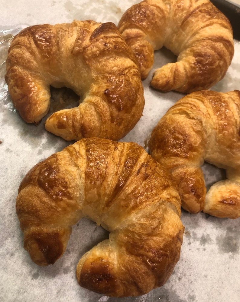 May include: Four golden brown croissants on a white parchment paper surface. The croissants are arranged in a semi-circle.