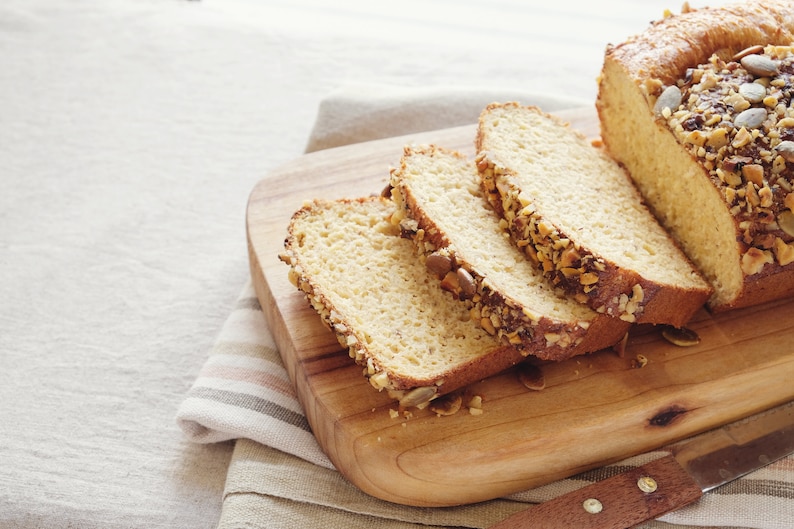 May include: A loaf of sliced bread with nuts and seeds on a wooden cutting board. The bread is light brown and the nuts and seeds are a variety of colors.
