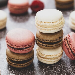May include: A stack of colorful macarons with pink, white, and brown frosting. The macarons are arranged on a dark brown surface with raspberries scattered around.