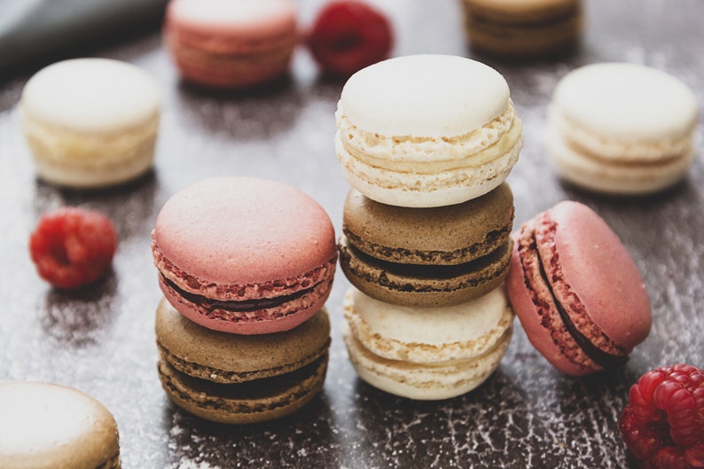 May include: A stack of colorful macarons with pink, white, and brown frosting. The macarons are arranged on a dark brown surface with raspberries scattered around.