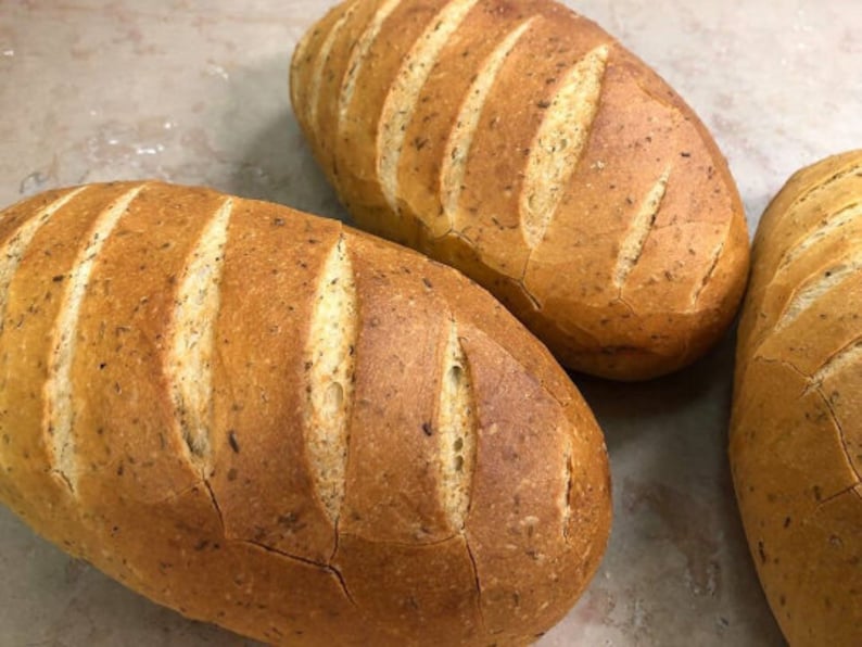 May include: Three loaves of freshly baked bread with a golden brown crust and a slightly cracked surface. The bread has a rustic appearance and is likely made with whole wheat flour.