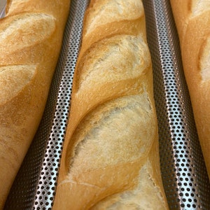 May include: Three long, golden brown baguettes are lined up on a silver baking sheet with a perforated surface. The baguettes are slightly browned and have a slightly crispy crust.