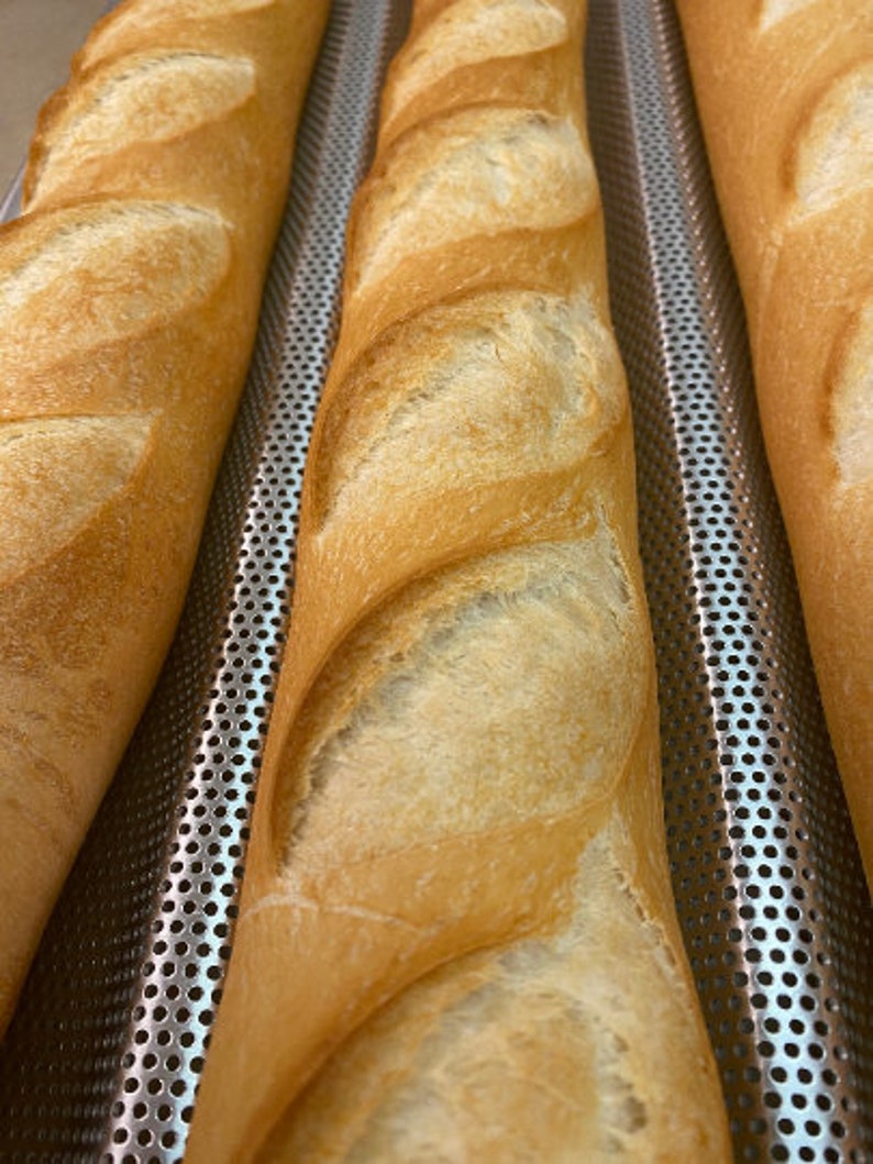 May include: Three long, golden brown baguettes are lined up on a silver baking sheet with a perforated surface. The baguettes are slightly browned and have a slightly crispy crust.