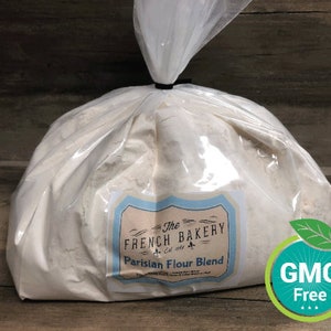 May include: A white plastic bag filled with white flour. The bag has a label that reads "The French Bakery Est. 2014 Parisian Flour Blend". A green and blue sticker with the text "GMO Free" is in the lower right corner of the image.