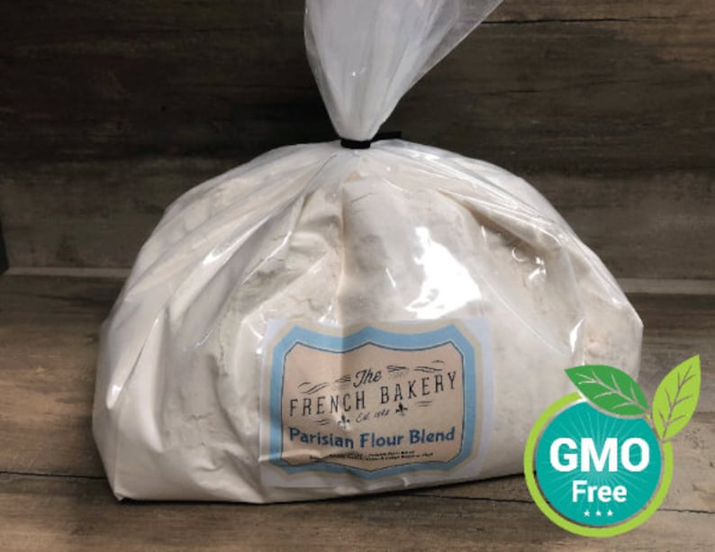 May include: A white plastic bag filled with white flour. The bag has a label that reads "The French Bakery Est. 2014 Parisian Flour Blend". A green and blue sticker with the text "GMO Free" is in the lower right corner of the image.