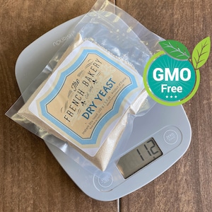 May include: A white scale with a digital display showing 11.2. A clear plastic bag containing a beige powder labeled "The French Bakery Dry Yeast" is on the scale. The bag is labeled with the text "Instant Dry Yeast 100 g/3.5 oz - Product of France" and "Best Use by 10/2021". A green and white sticker with the text "GMO Free***" is in the upper right corner of the image.