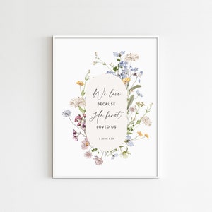 May include: Framed art print featuring the quote "We love because He first loved us" surrounded by watercolor floral illustrations in shades of pink, blue, yellow, and white. The artwork is set against a white background.