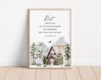 May include: Framed art print featuring a watercolor illustration of a cabin in a mountain landscape. The print includes the text "Rest, come to me... and I will give you rest." from Matthew 11:28. The frame is a light brown color.