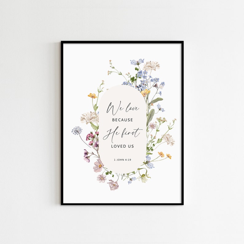 May include: Framed art print featuring the quote "We love because He first loved us" surrounded by a floral border. The flowers are in shades of pink, yellow, blue, and white, set against a white background. The print is in a black frame.