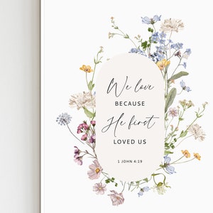 May include: A floral print art print with the text "We love because He first loved us" and the Bible verse 1 John 4:19. The print features a variety of flowers in shades of pink, yellow, blue, and white.