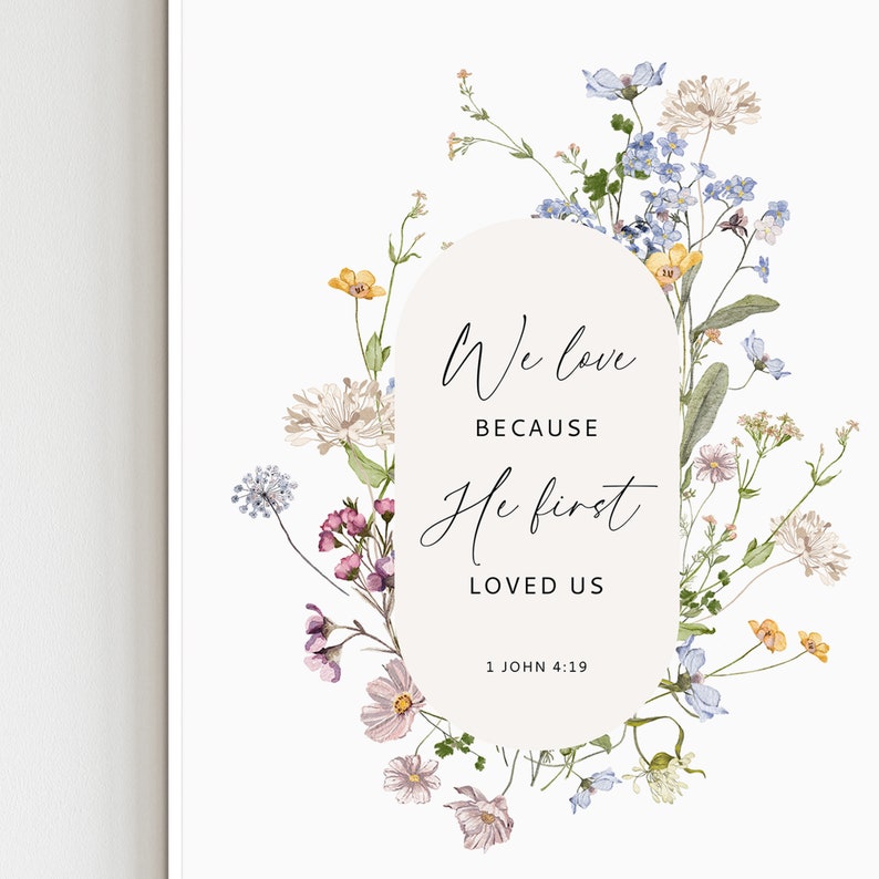 May include: A floral print art print with the text "We love because He first loved us" and the Bible verse 1 John 4:19. The print features a variety of flowers in shades of pink, yellow, blue, and white.