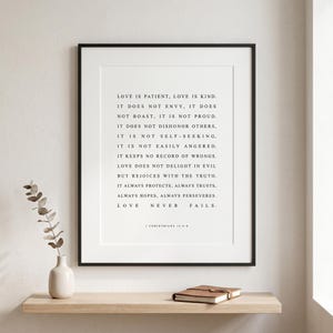 May include: A framed wall art print featuring the text of 1 Corinthians 13:4-8 in black serif font on a white background. The text reads: Love is patient, love is kind. It does not envy, it does not boast, it is not proud. It does not dishonor others.