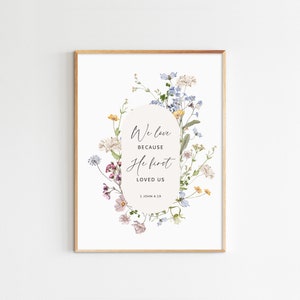 May include: A framed art print featuring a floral design with the text "We love because He first loved us" from 1 John 4:19. The artwork has a white background with a gold frame and a soft, watercolor style.