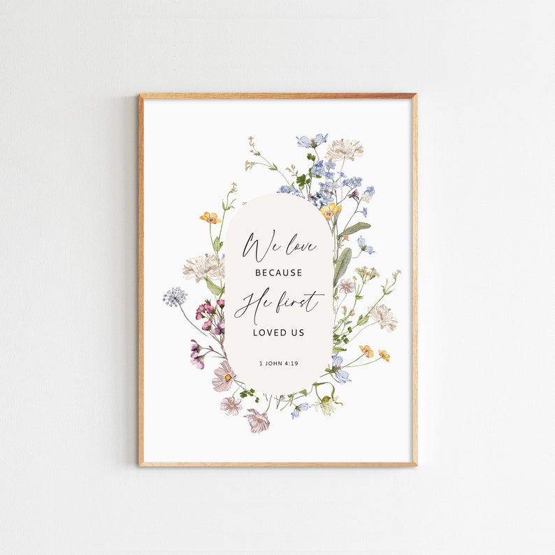 May include: A framed art print featuring a floral design with the text "We love because He first loved us" from 1 John 4:19. The artwork has a white background with a gold frame and a soft, watercolor style.