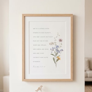 Puede incluir: Una impresión de arte mural enmarcada que presenta el texto 'She is clothed with strength and dignity, and she laughs without fear of the future. When she speaks, her words are wise, and she gives instructions with kindness. Proverbs 31:25-26' junto a una ilustración floral de flores silvestres azules, amarillas y rosas en un marco de madera clara.