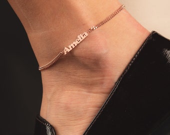Puede incluir: Un brazalete de tobillo de oro rosa con el nombre "Amelia" en él. El brazalete está en el tobillo de una persona, y lleva zapatos negros.