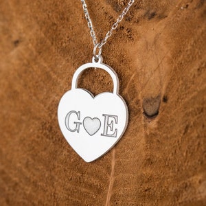 Puede incluir: Collar de plata con colgante en forma de corazón con diseño de candado. El colgante tiene las letras "G" y "E" grabadas, con un pequeño corazón entre ellas.