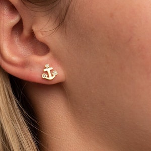 May include: Gold anchor earrings with small clear gemstones. The earrings are a simple and elegant design.