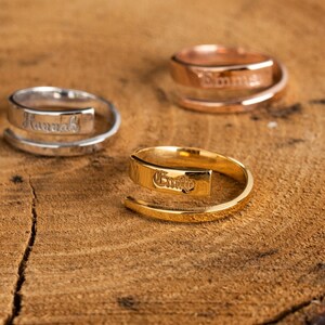 May include: Three personalised rings on a wooden surface. The rings are made of silver, gold, and rose gold. The names "Hannah", "Emily", and "Emma" are engraved on the rings.