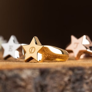 Puede incluir: Tres anillos de oro en forma de estrella con diferentes iniciales grabadas. Los anillos están sobre una superficie de madera.
