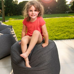 Puede incluir: Una niña pequeña con una camisa rosa se sienta en un puf gris en un patio trasero con césped.