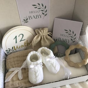 May include: A baby gift set in a box, including white knitted baby booties with bows, a beige muslin blanket, and a round milestone card that says "12 Months". The box also contains a small drawstring bag, two cards that say "Hello Baby", and two baby teethers.