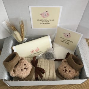 Pode incluir: Uma caixa de presente para bebês com vários itens. Inclui cartões com uma ilustração de urso e o texto "CONGRATULATIONS ON YOUR BABY NEWS". Há também meias de urso de pelúcia, uma musselina e um pequeno item com o texto "FUEL FOR MUM & DAD".