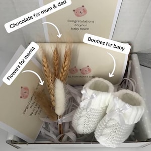 Op de afbeelding: Een geschenkdoos met witte gebreide babyslofjes, wenskaarten en tarwearen. De kaarten hebben een berenillustratie en de tekst "Gefeliciteerd met je babynieuws!". De slofjes hebben witte strikken.
